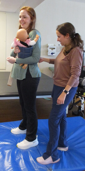 Patient is practicing standing while holding a lifesize babydoll with her occupational therapist beside her