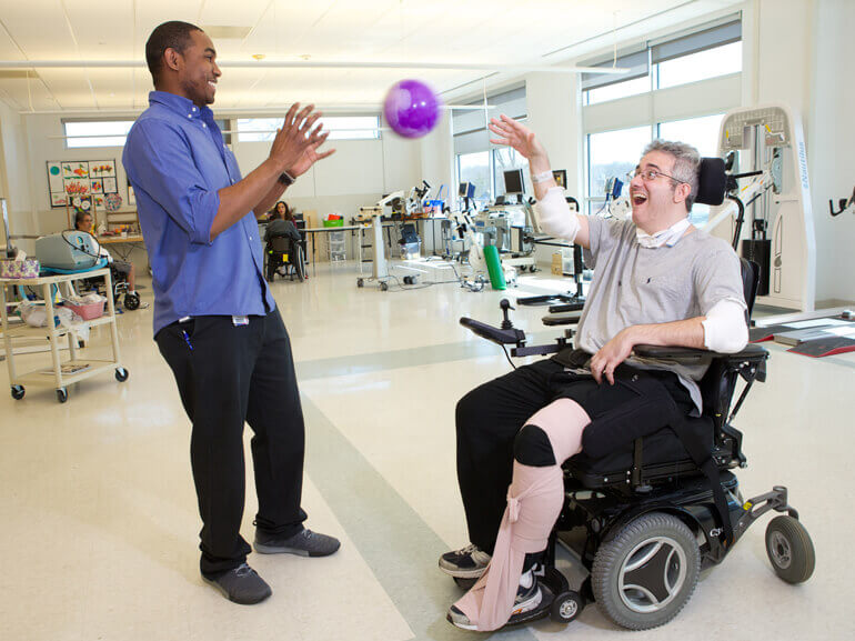 Male patient in wheelchair practicing throwing and catching a ball with a physical therapist