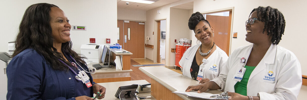 A group of three nurses gathered at a hospital nurse's station talking.
