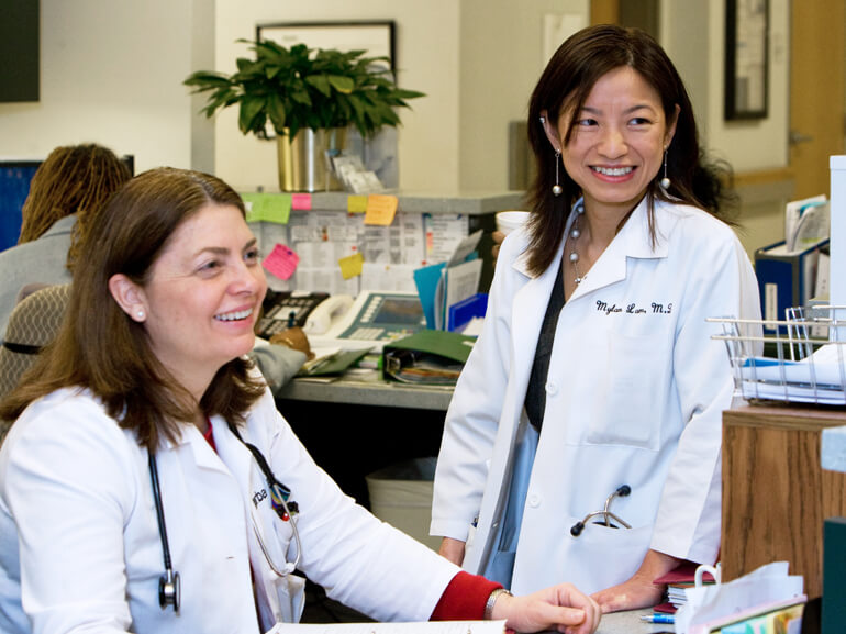 Two female doctors in white coats at a nurse's station in a hospital.