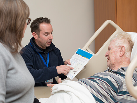 Male nurse explaining a document to a senior-aged male patient lying in a hospital bed.