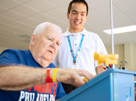 White haired man putting small items into a plastic bin on a table.