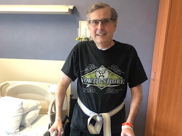 Man wearing Northshore logo black t-shirt standing with a walker in a hospital room.