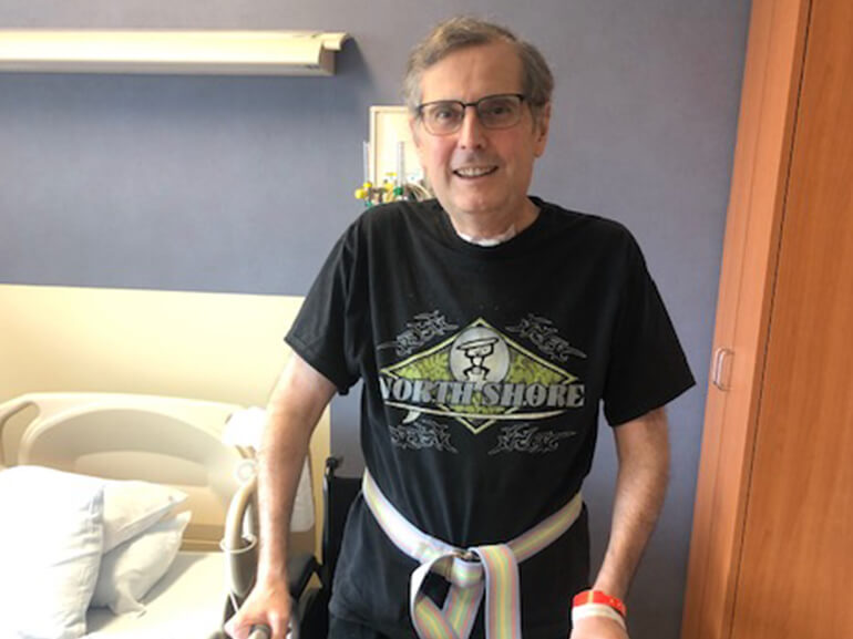James wearing a Northshore logo black t-shirt standing with a walker in a hospital room.