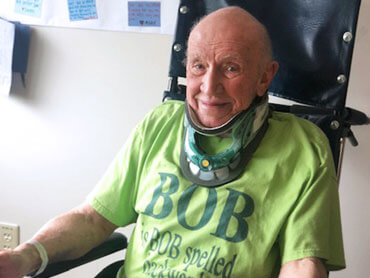 Man in a green t-shirt wearing a neck brace sitting in a hospital room.