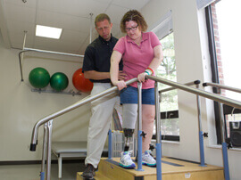Male therapist spotting female patient with leg prosthesis as she climbs a stair.