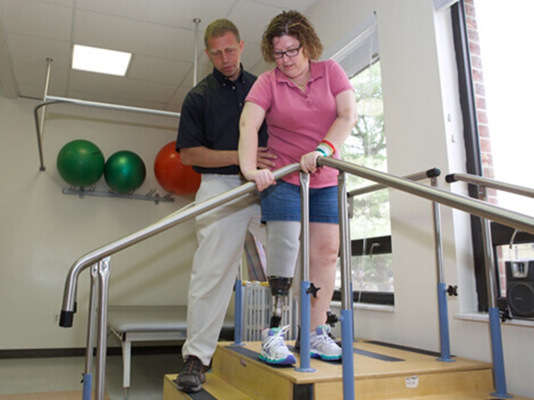 Woman with prosthetic leg being helped by a therapist to climb a stair in a gym.