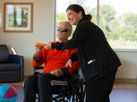Dark-haired female therapist leaning toward male patient sitting in a wheelchair and wearing dark glasses.