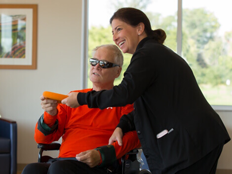 Dark-haired female therapist leaning toward man in wheelchair wearing dark glasses.