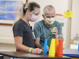 Therapist standing next to a man stacking colorful plastic tubes together for hand therapy exercise.