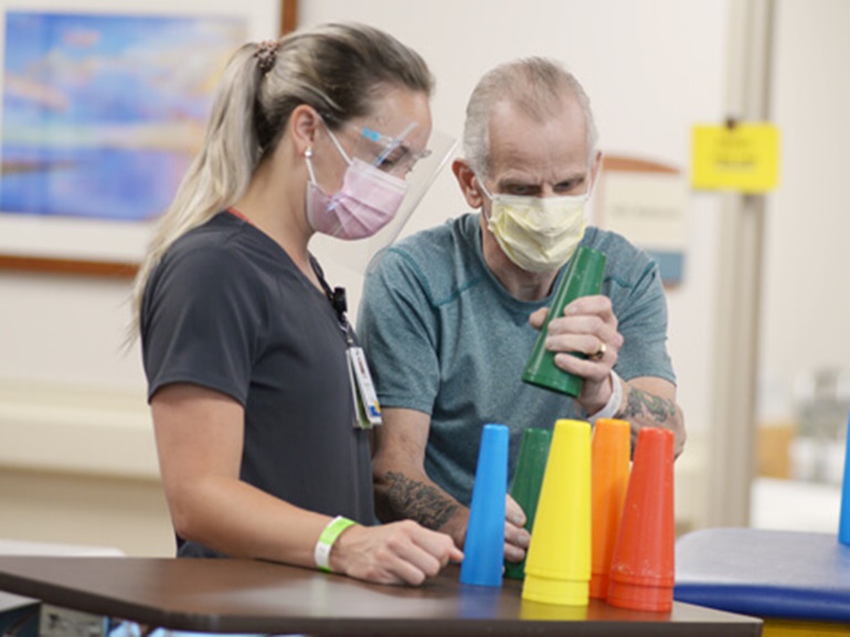A male patient wearing a hospital mask and stacking small colored plastic cones as part of a therapy exercise.