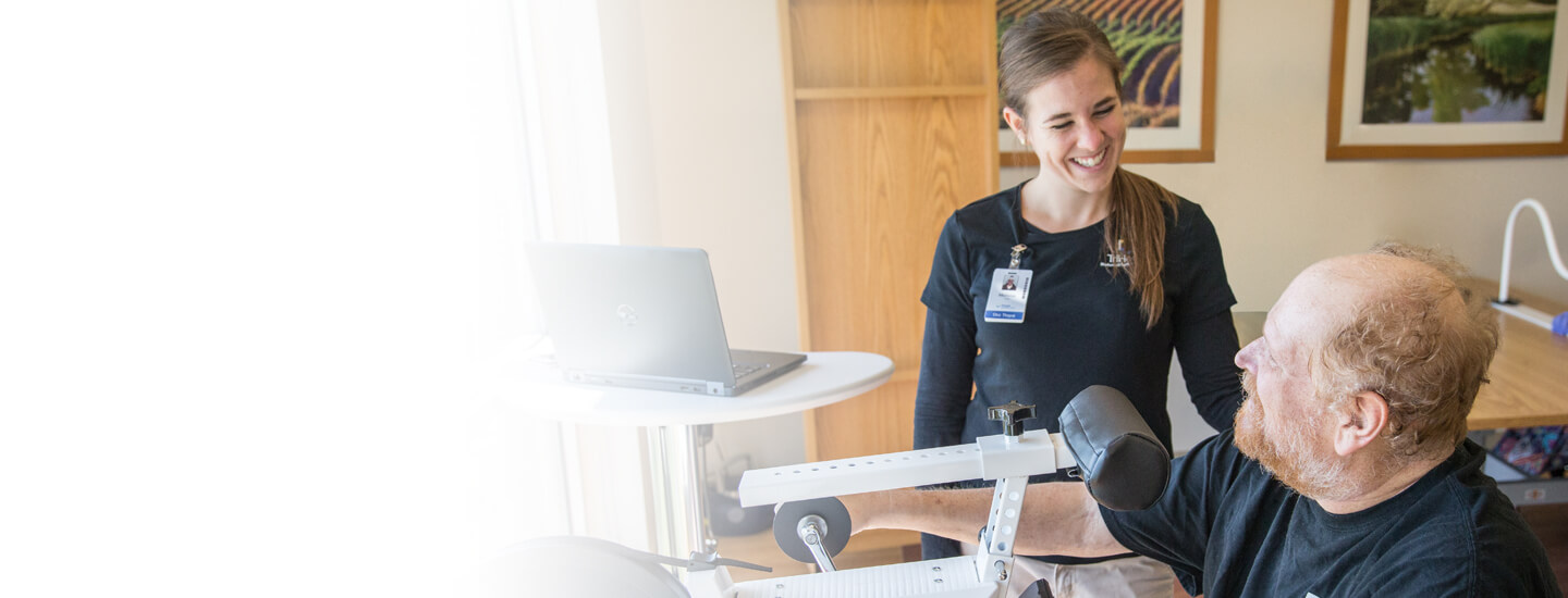 Young female therapist smiling at a man using an arm pedal exerciser in a gym.